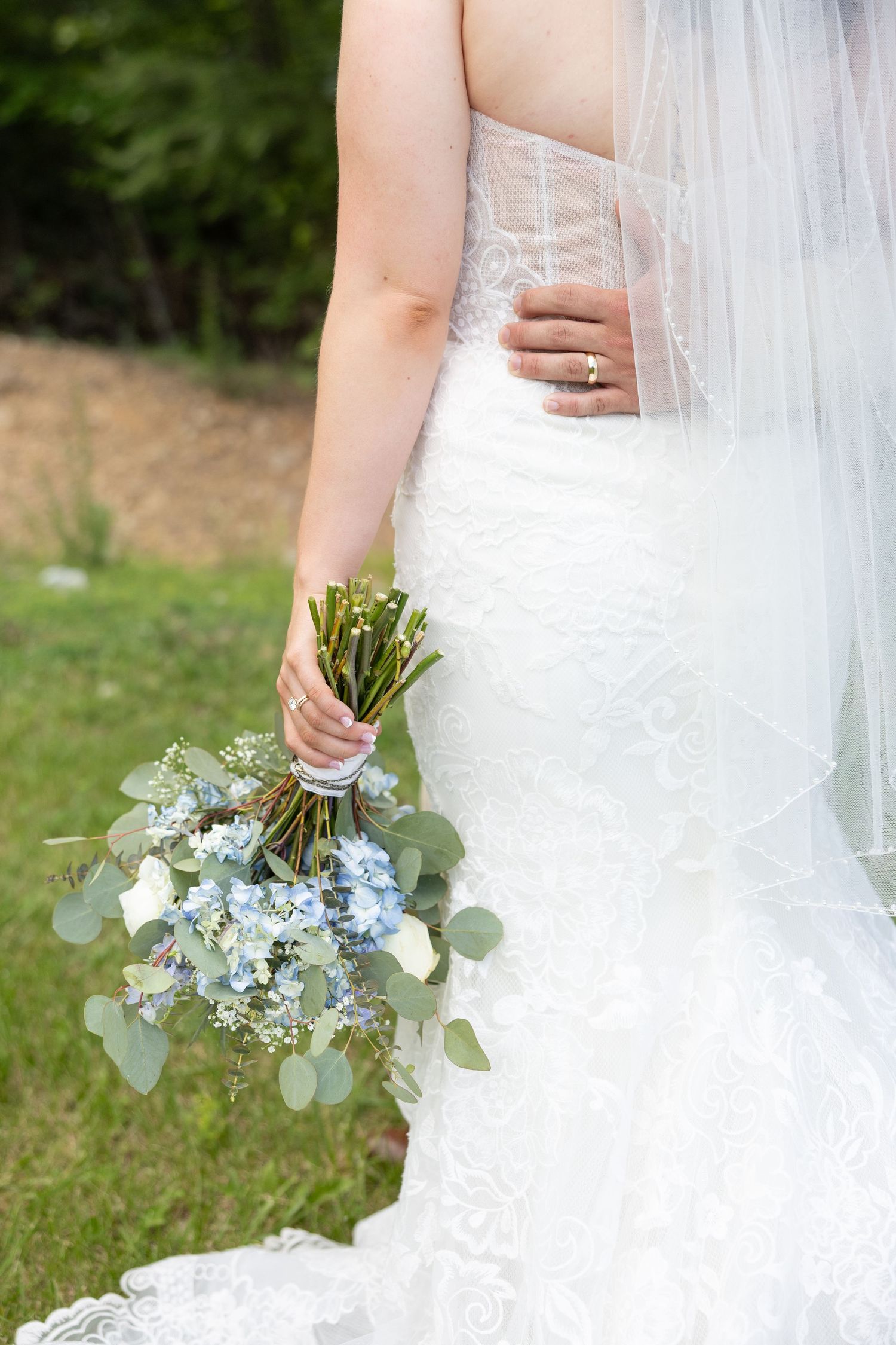 Detail of bride holding a delicate blue and white bouquet with greenery against her lace wedding dress and veil.