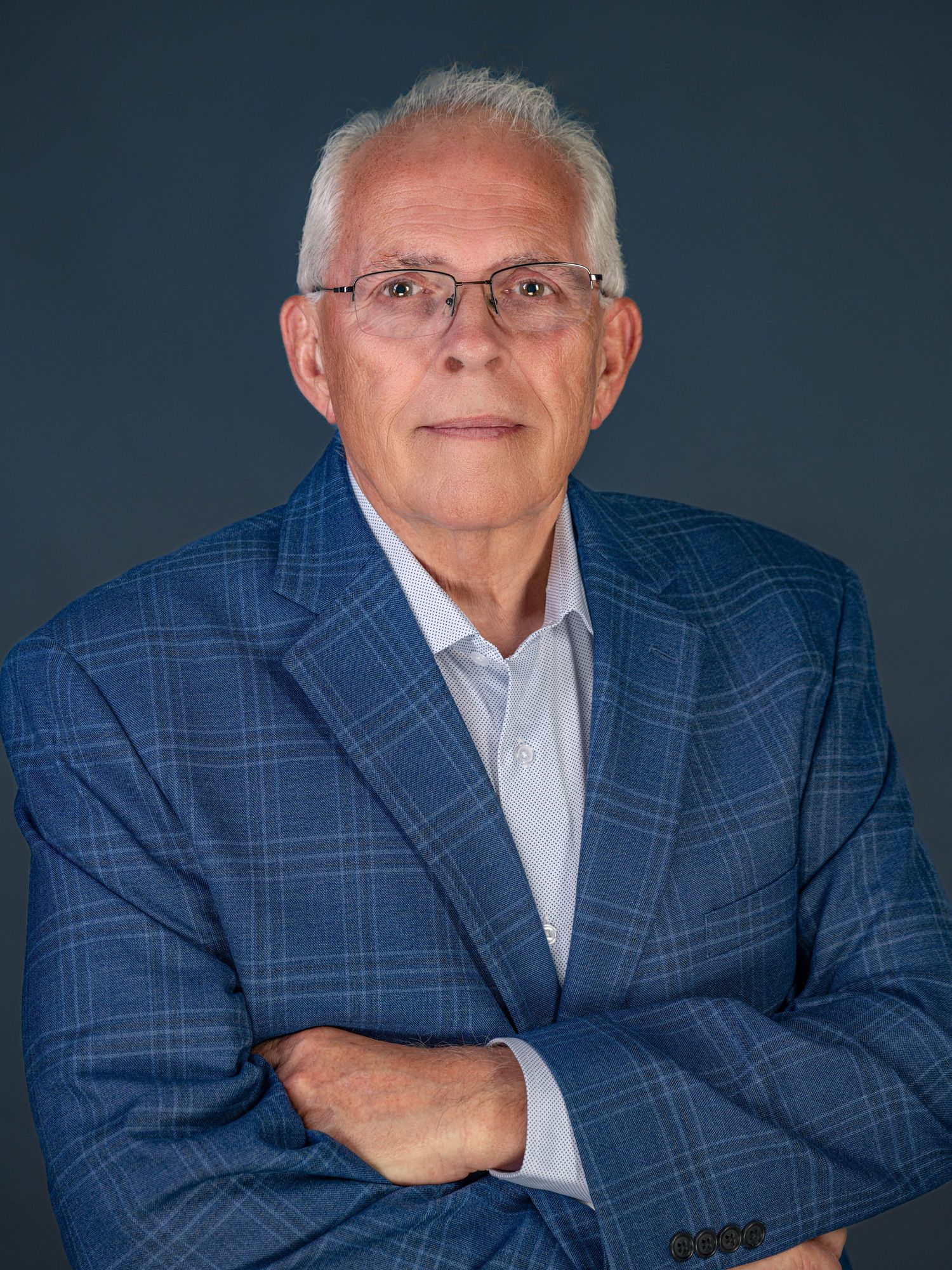 Professional studio portrait of a senior person in a blue plaid blazer with glasses against a dark background.