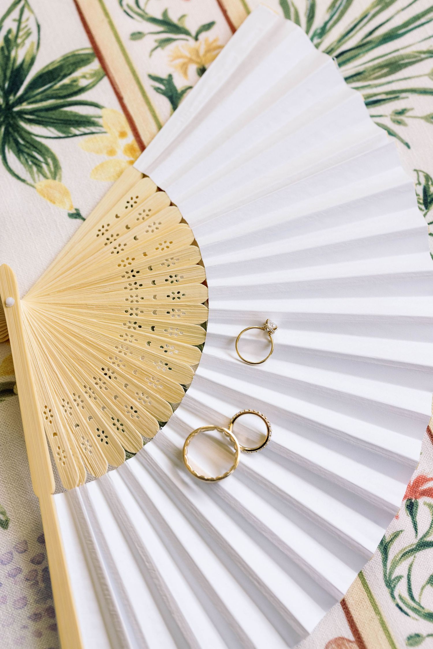 White paper fan with gold bamboo ribs displays two gold wedding rings against a floral background.