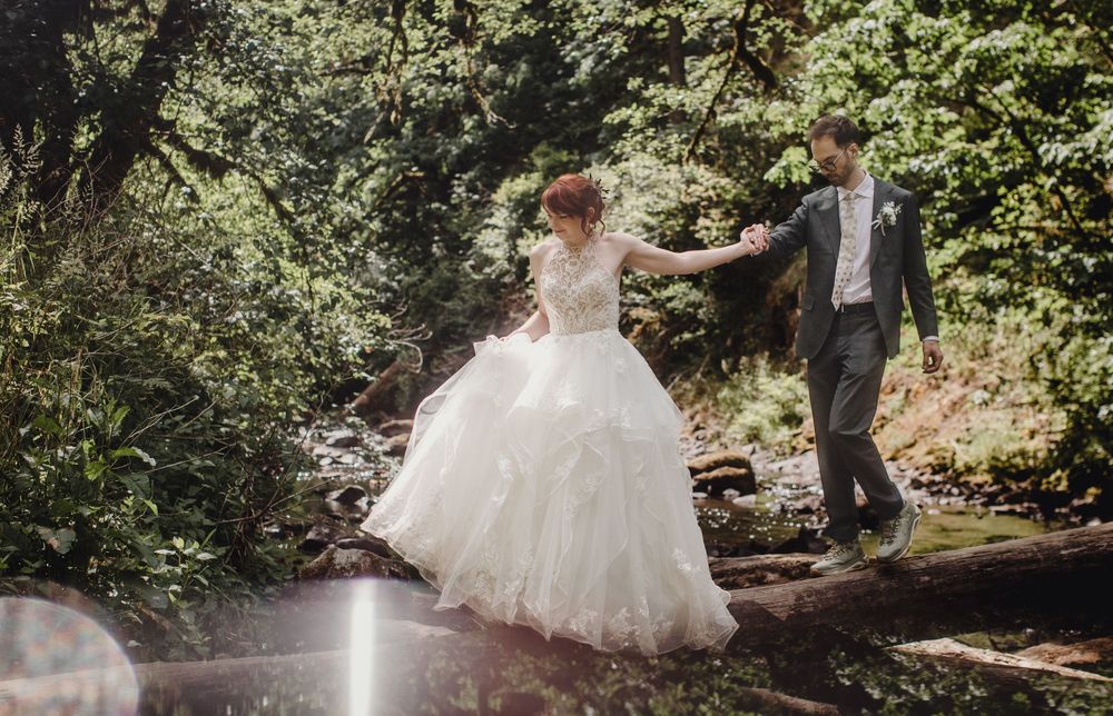 A bride in a white ballgown and groom walking along a scenic wooded path by a stream on their wedding day.