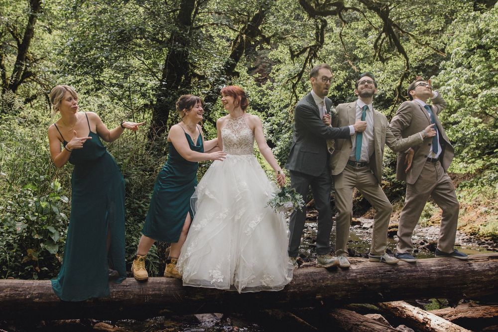 Wedding party stands balanced on fallen log in lush green forest setting during outdoor celebration.