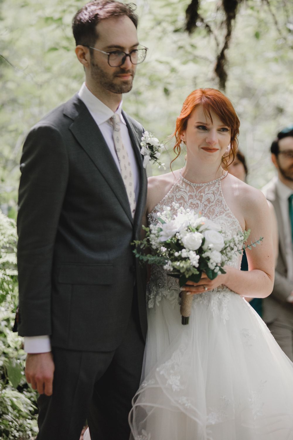 A bride in a lace wedding gown holds a white bouquet next to a groom in a dark suit outdoors.