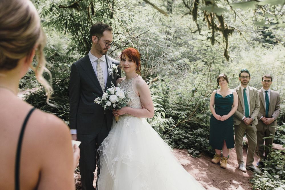 Elegant outdoor wedding ceremony in a lush forest setting with groomsmen and bridesmaids looking on in warm sunlight.