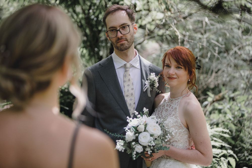 A couple at their woodland wedding ceremony poses with white floral bouquet while listening to officiant.