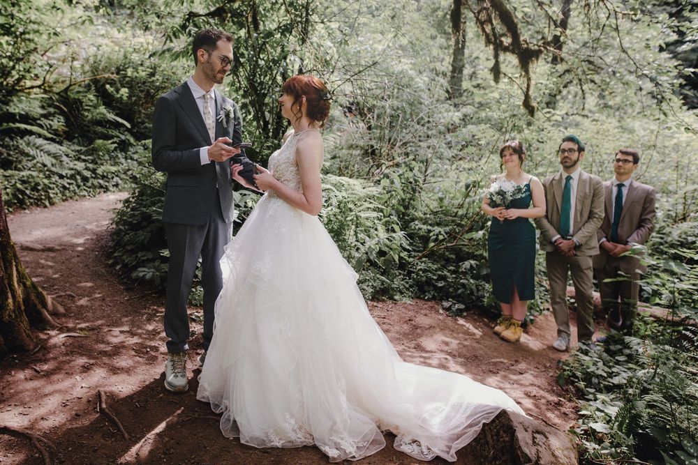 Wedding party poses together on wooded forest path surrounded by lush greenery and sunlight filtering through trees.