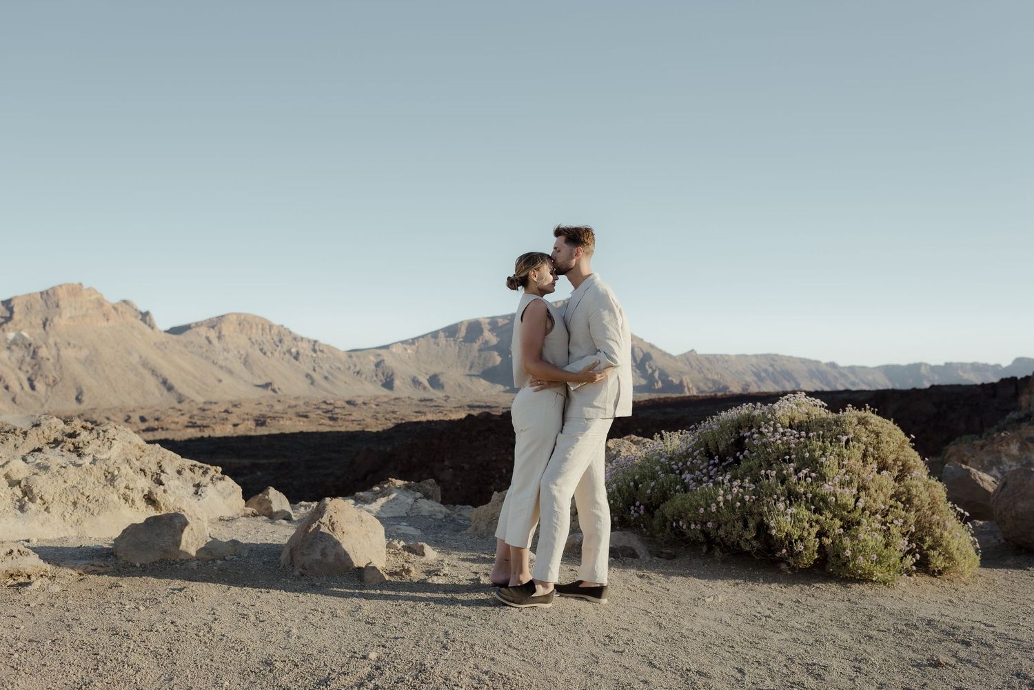 A couple embraces while standing on rocky desert terrain with mountains in the background during sunset.