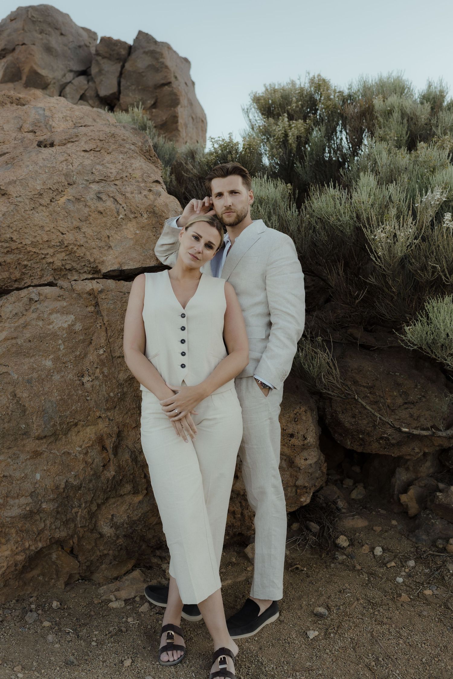Couple in white outfits share an intimate moment against a desert landscape with rocks and sage brush.