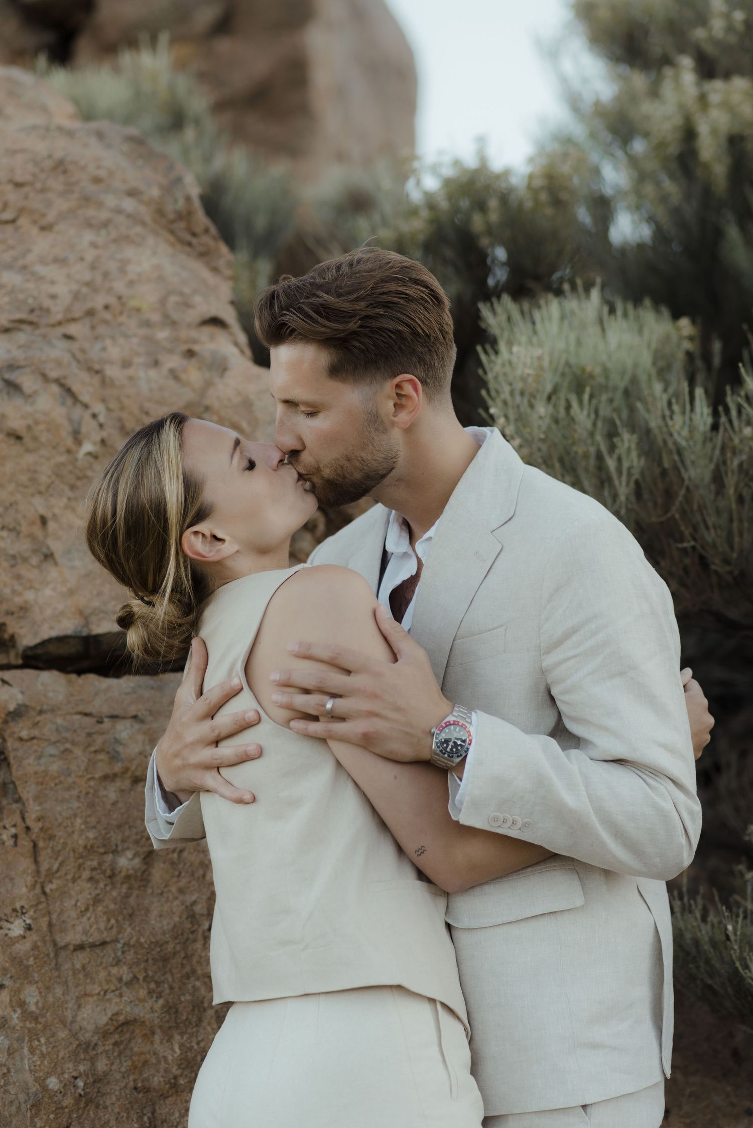 A romantic couple in beige formal attire share a tender embrace and kiss against a natural desert landscape backdrop.