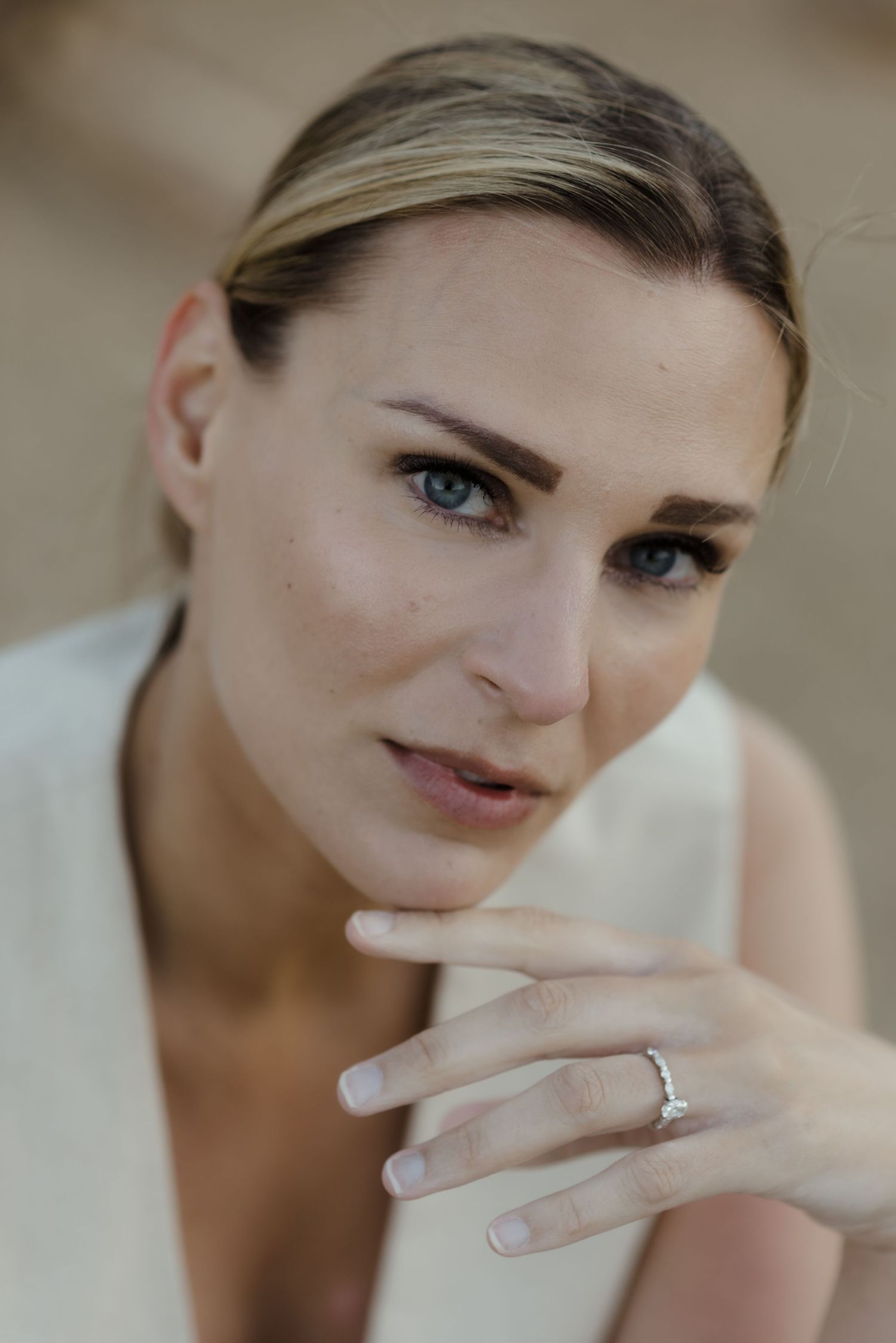 A close-up portrait of a person wearing minimal makeup with blue eyes and an engagement ring.