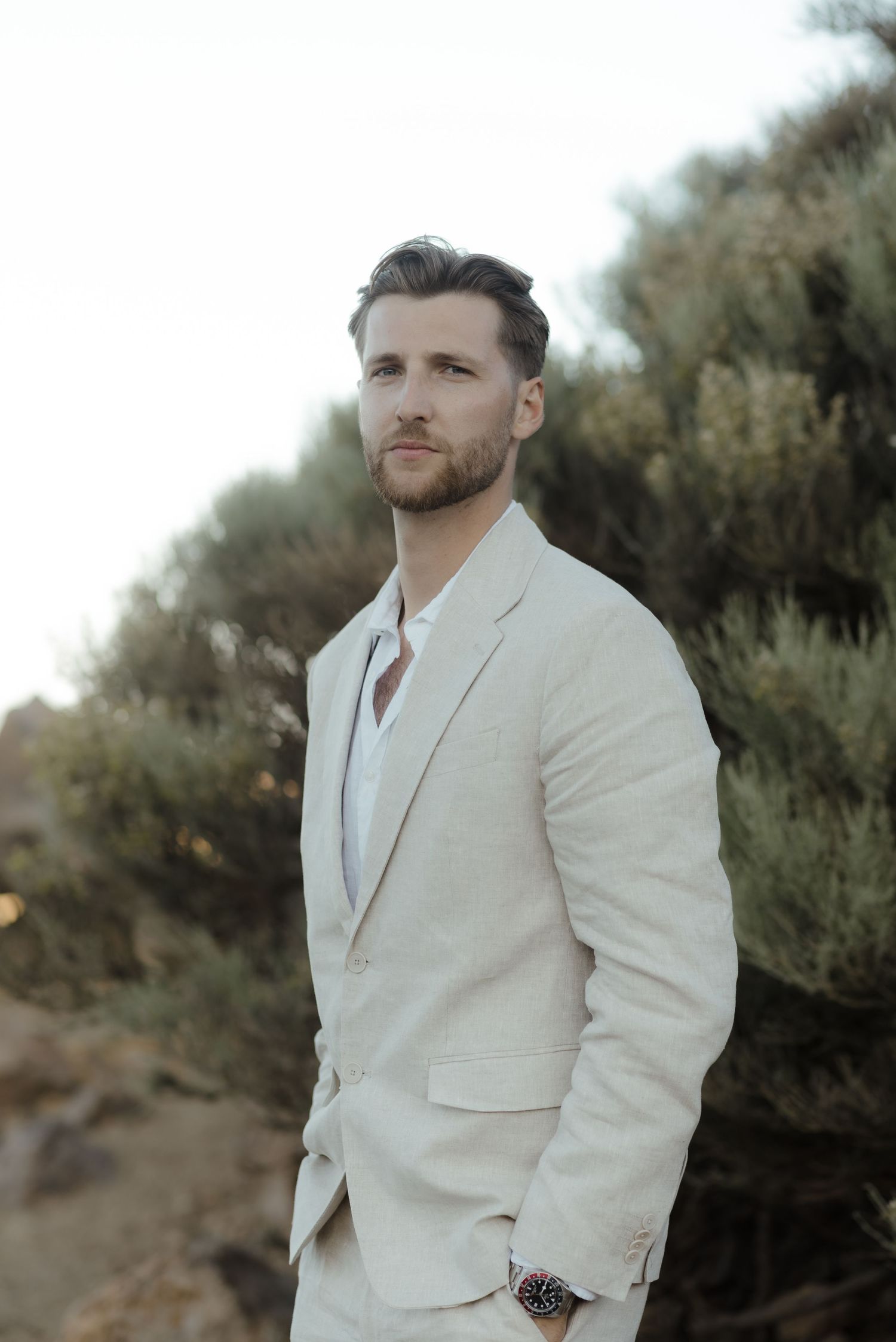 A man wearing a light beige suit jacket poses outdoors against a natural backdrop during golden hour.