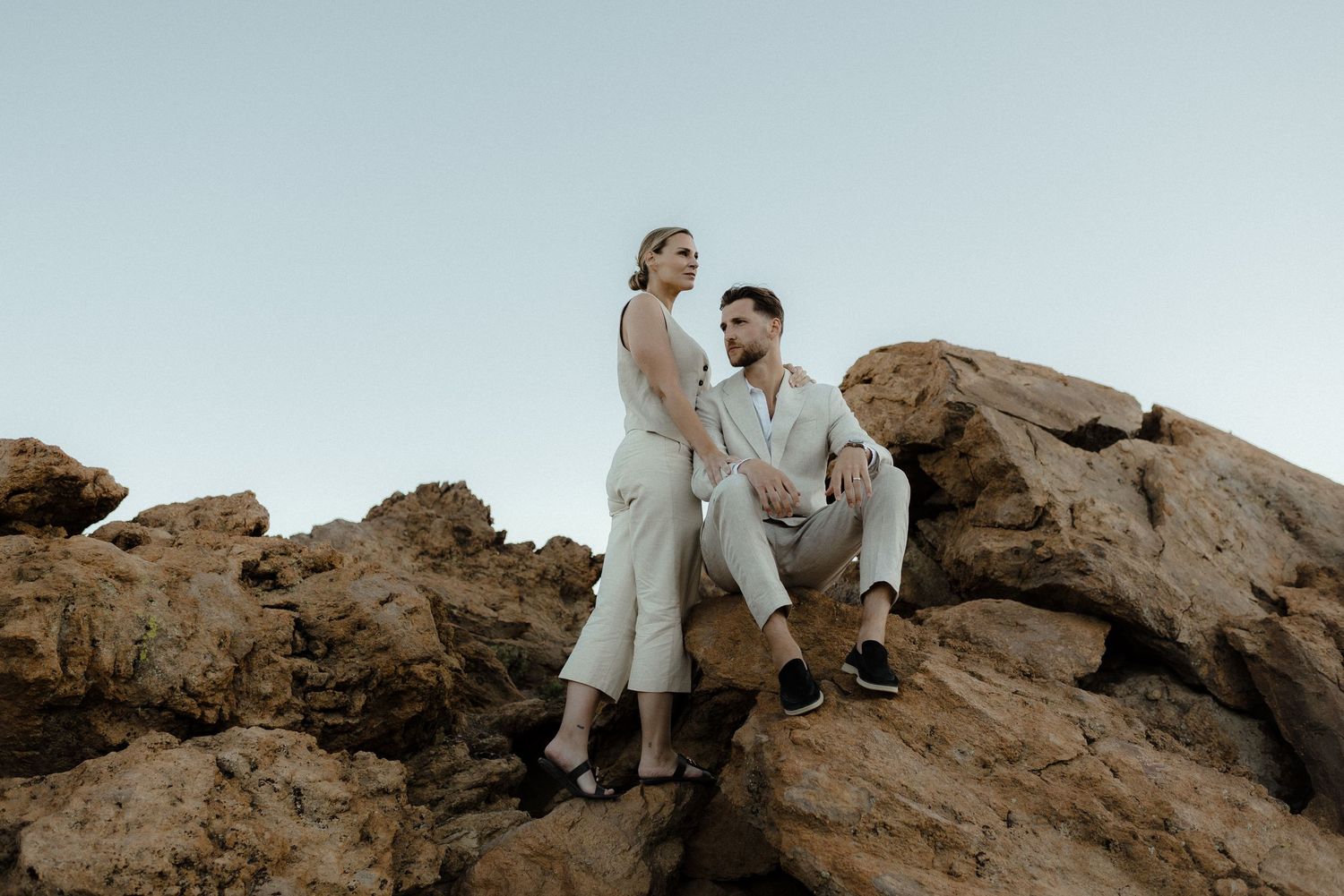 A couple in light-colored outfits poses together on rocky coastal cliffs at sunset for an engagement photoshoot.