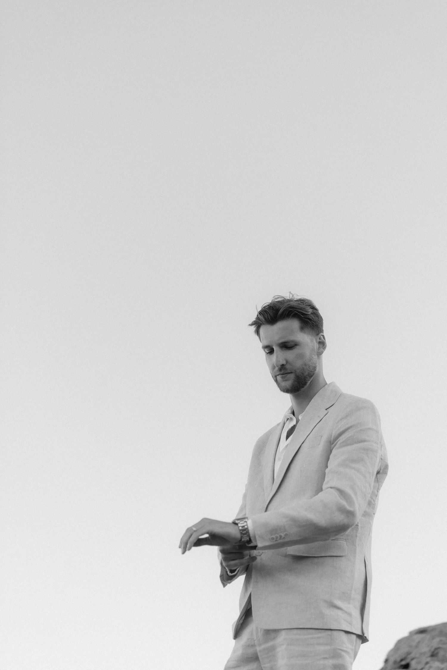 A figure in a light colored suit checks their watch against a bright sky background.