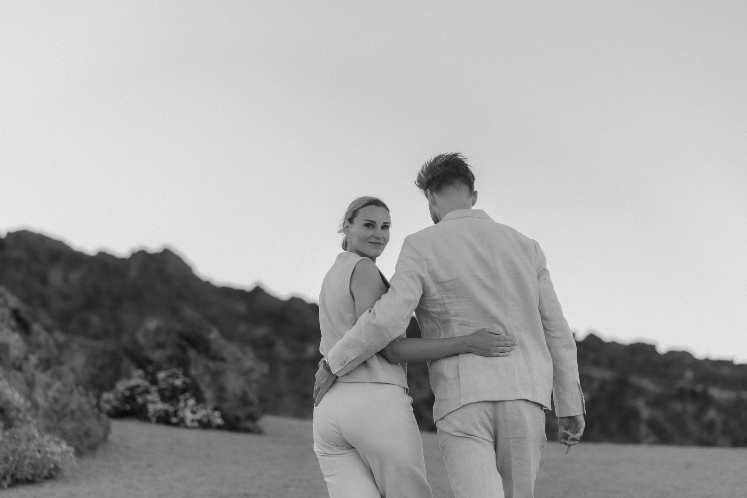 A couple walking together in light colored outfits along a hillside path.