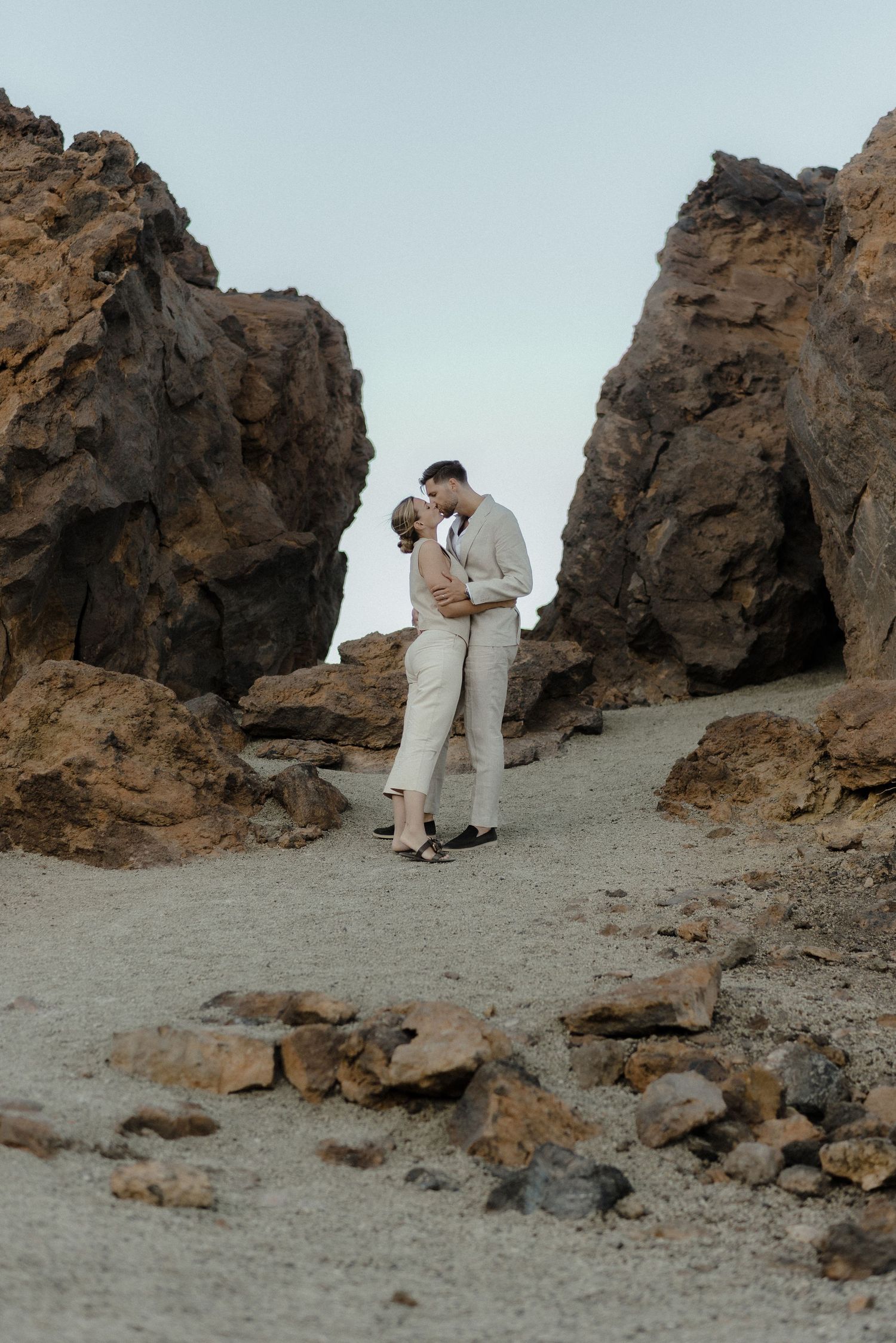 A couple shares a romantic moment while standing between towering rock formations on a dusty mountain trail.
