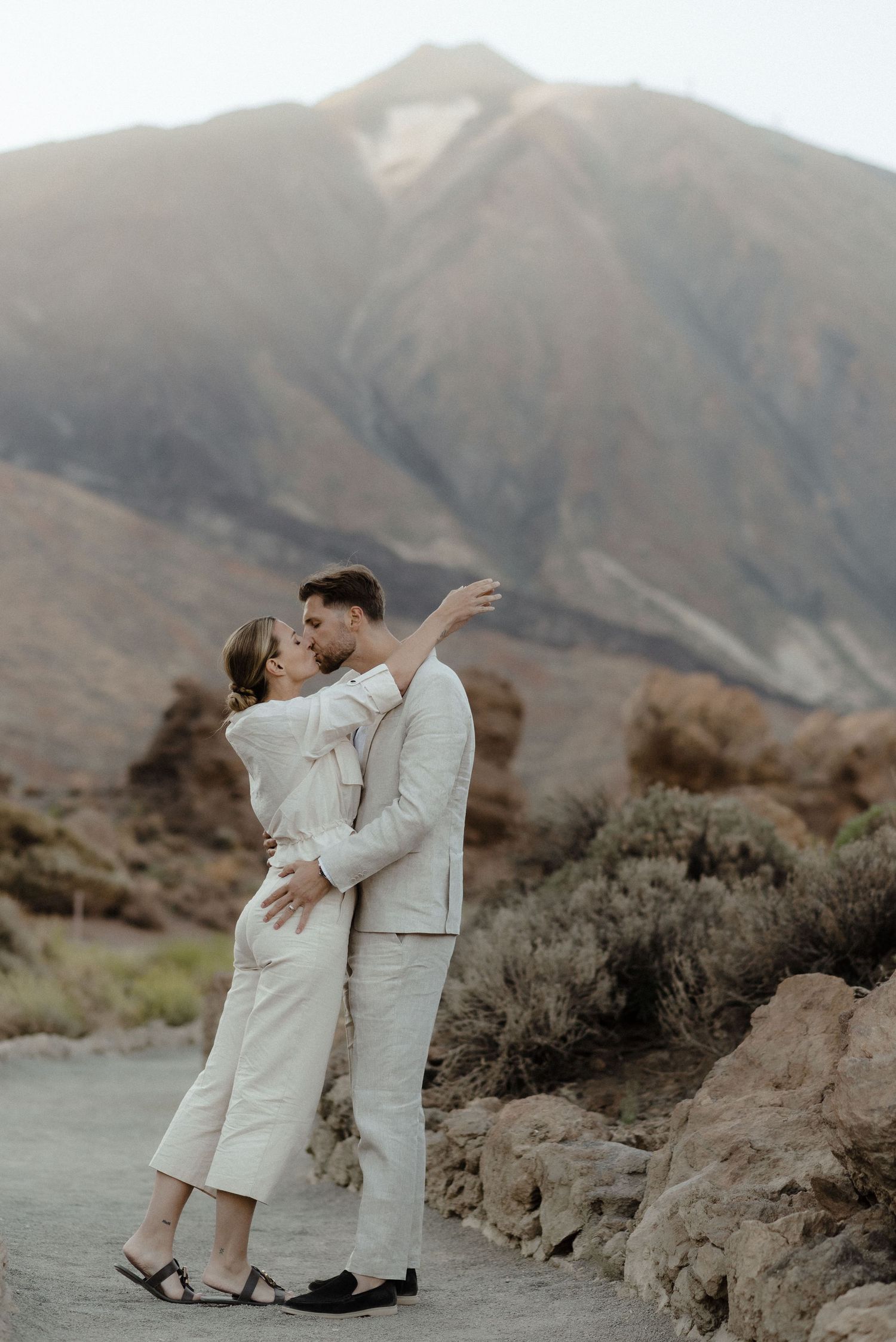 A romantic couple in white outfits embrace against a stunning mountain desert landscape.