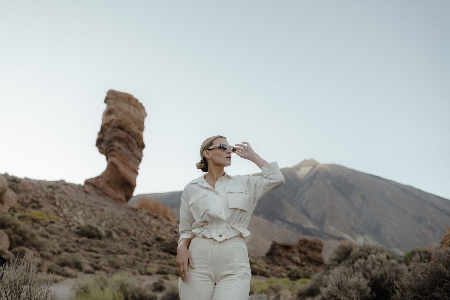 A woman in light clothing poses near a unique rock formation in a desert landscape.
