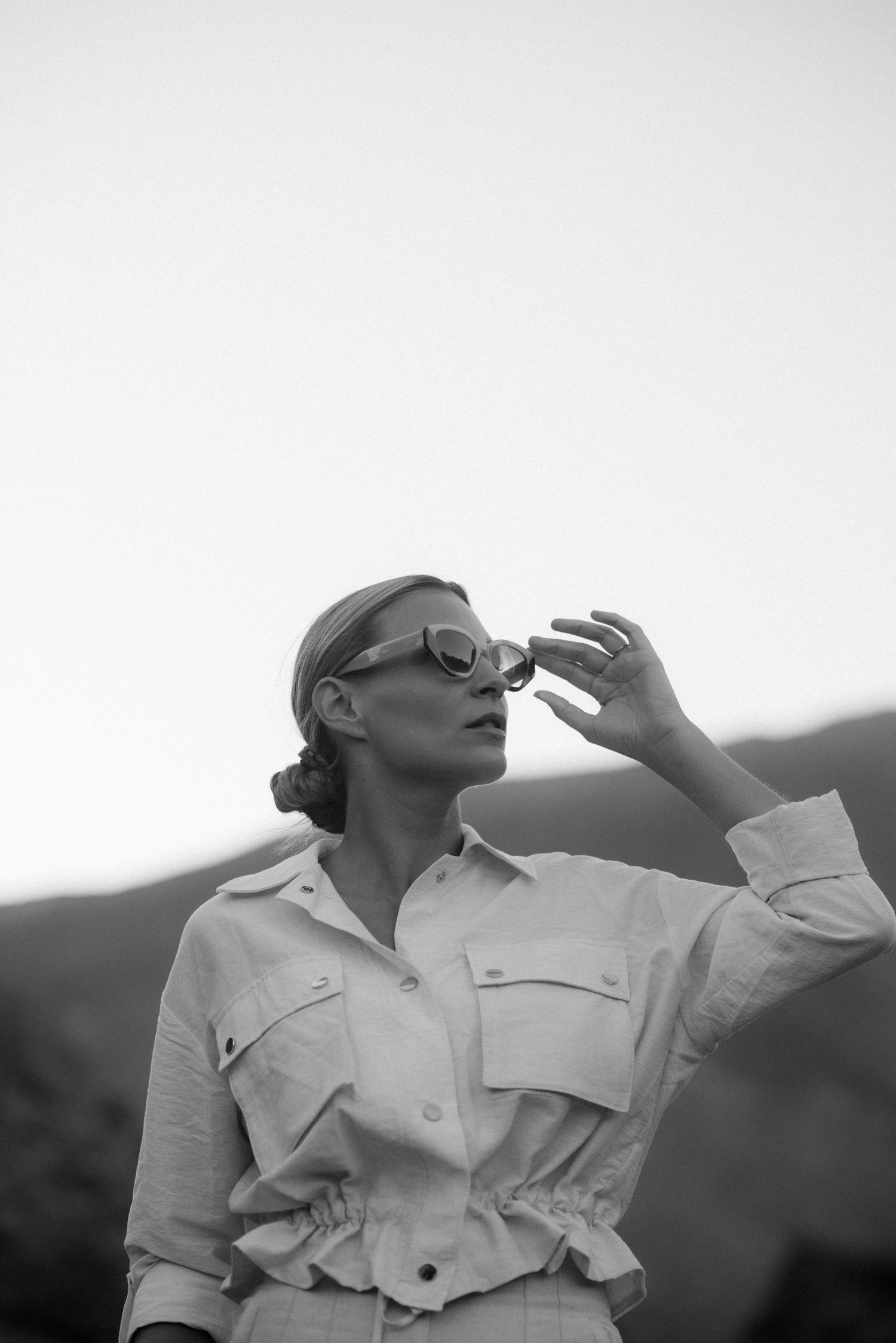 Artistic black and white portrait of a woman in sunglasses and light colored shirt against mountains.