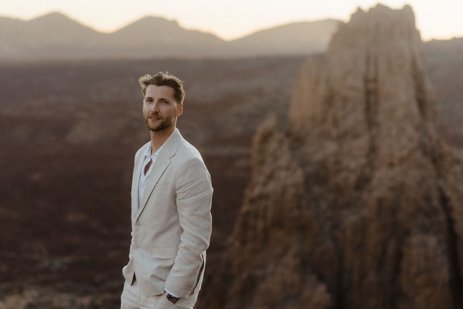 A person in a light suit stands on clifftop overlooking dramatic desert canyon landscape at sunset.