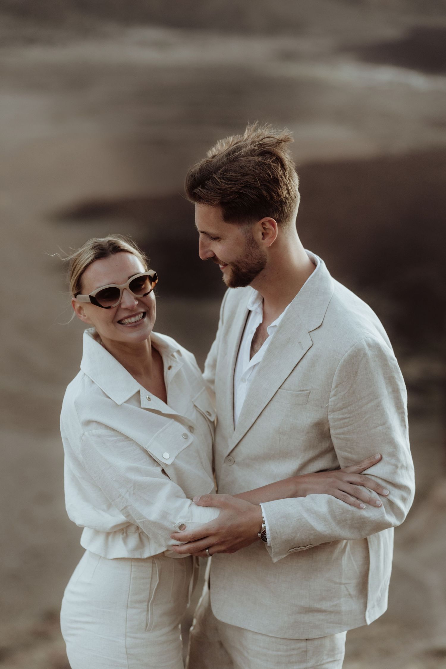 Romantic couple in white linen outfits share a tender moment against a moody desert landscape.