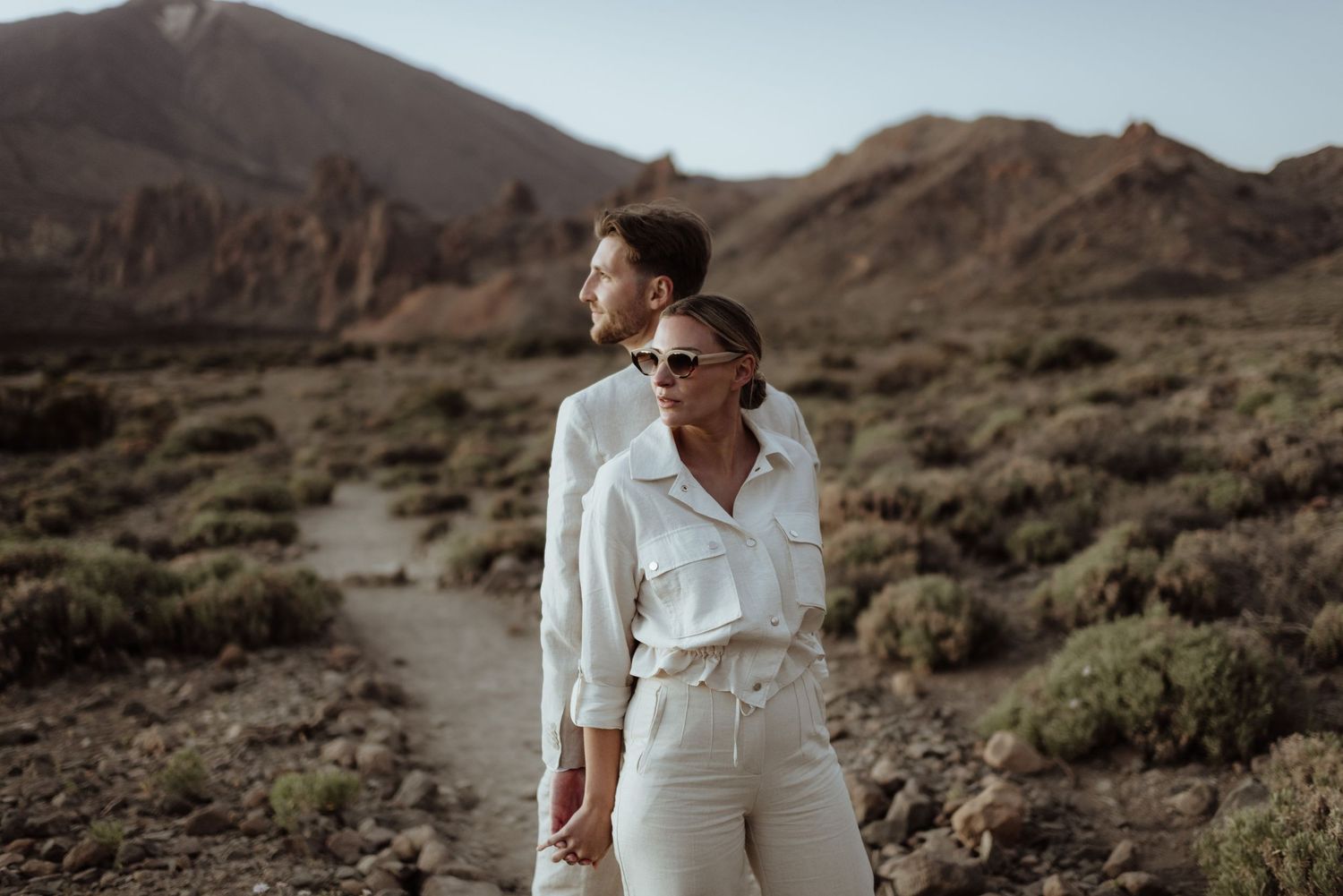 A couple in white outfits stands together against a mountainous desert backdrop.