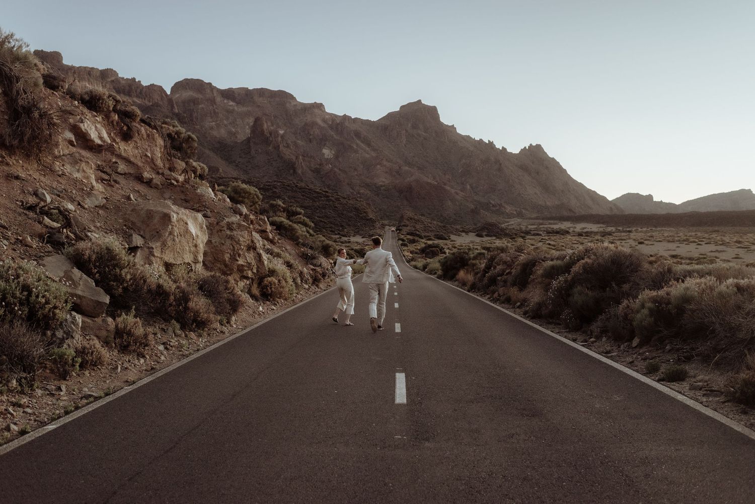 Two figures walk down an empty mountain road surrounded by dramatic rocky terrain.
