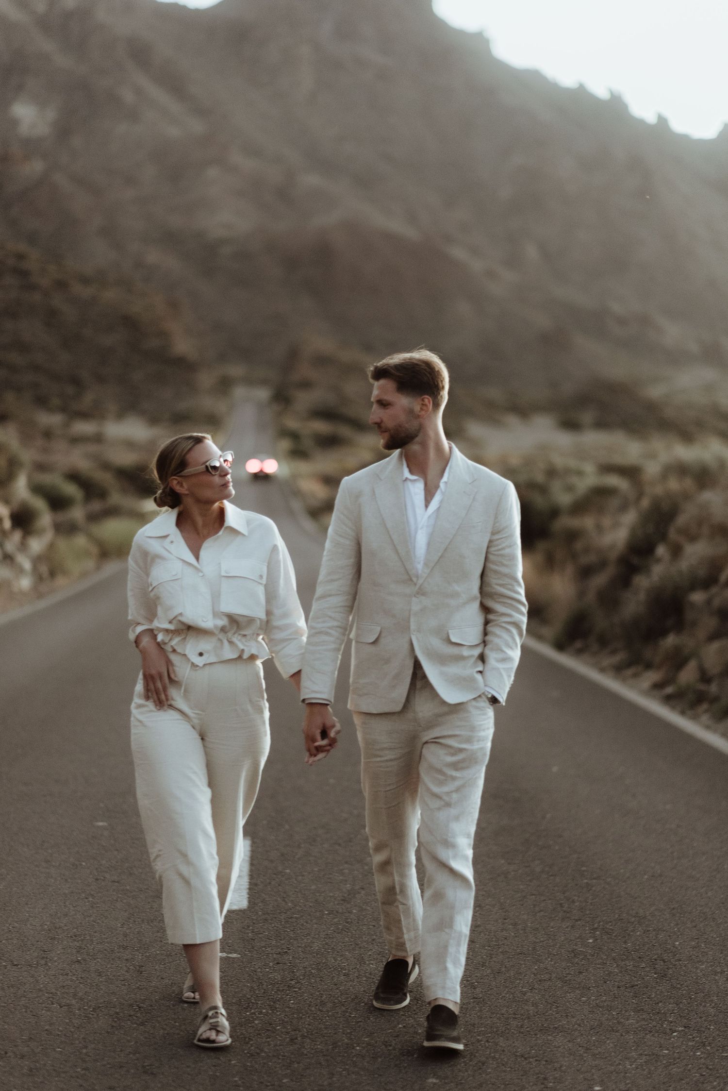 A couple in matching white outfits walks hand in hand down a desert road with mountains in the background.