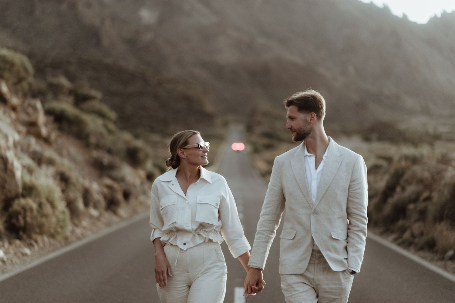 A couple in coordinated white outfits walking hand in hand along a desert mountain road.