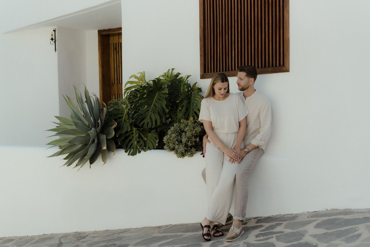 A couple stands against a white wall with tropical plants, dressed in light colored outfits for an engagement shoot.