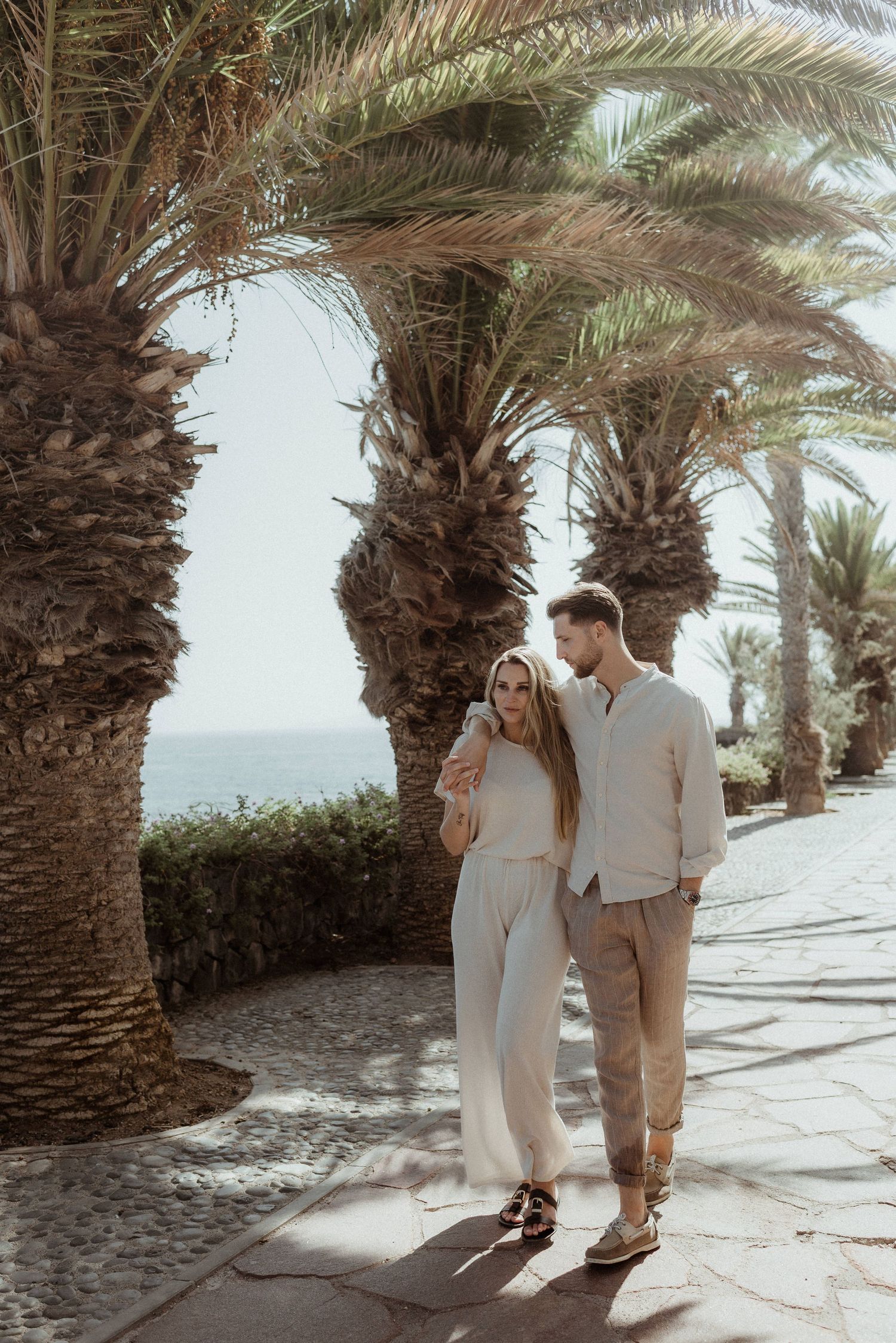 Couple walking together along a palm-lined path at a tropical resort under bright sunlight.