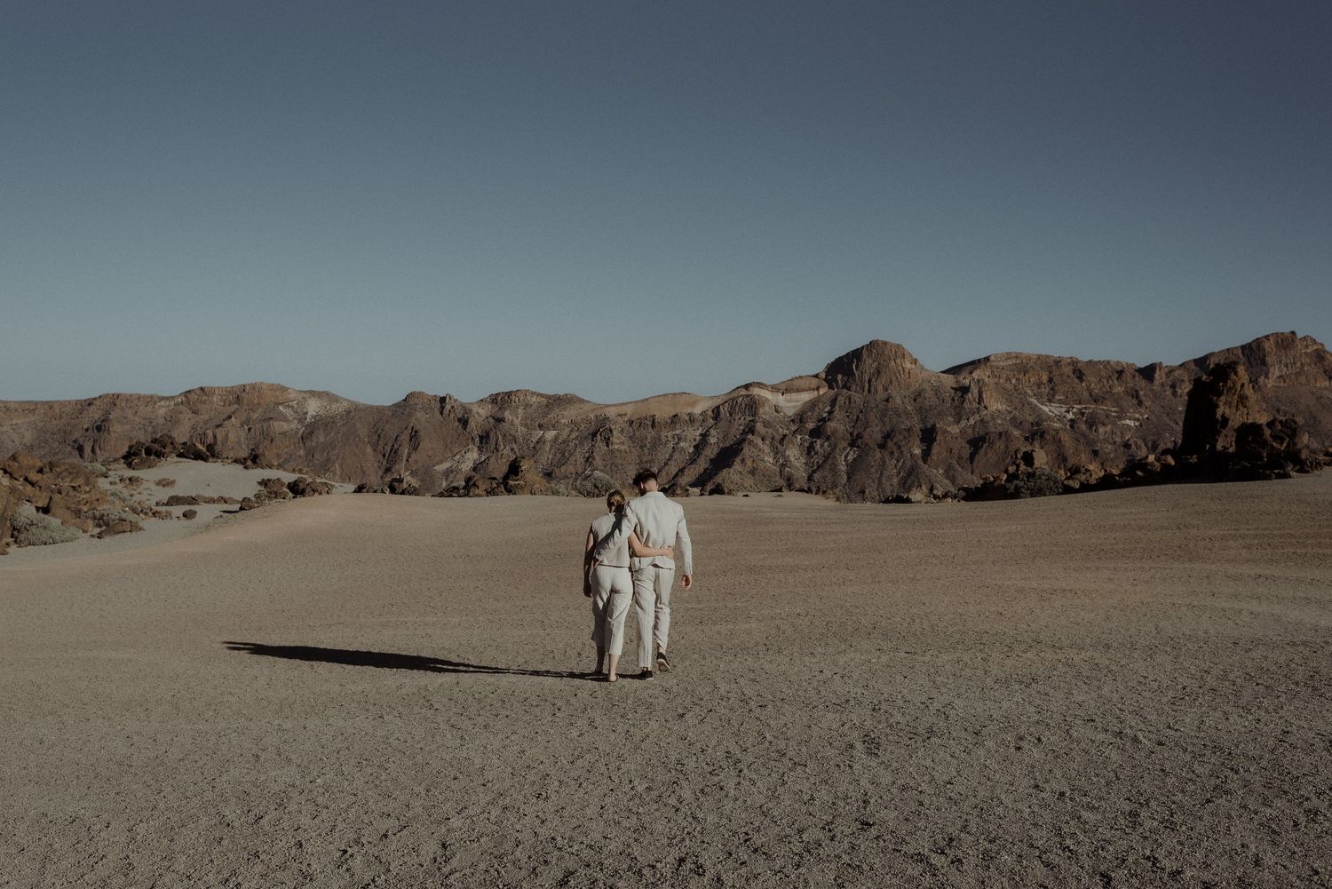 Two people walking hand in hand through a dramatic desert landscape with rocky mountains.