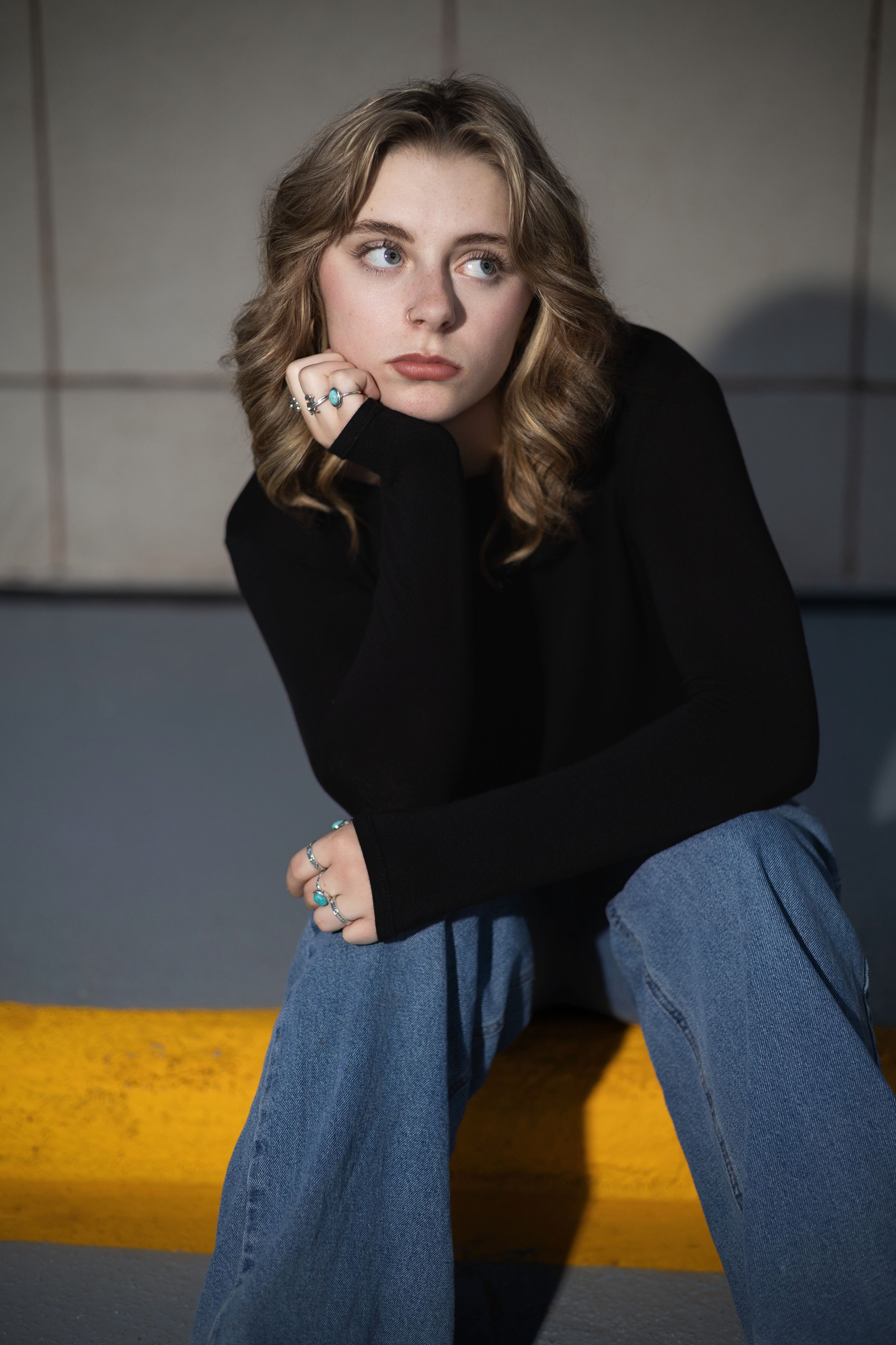 Person in black sweater and jeans sits pensively against blue wall with dramatic yellow lighting.