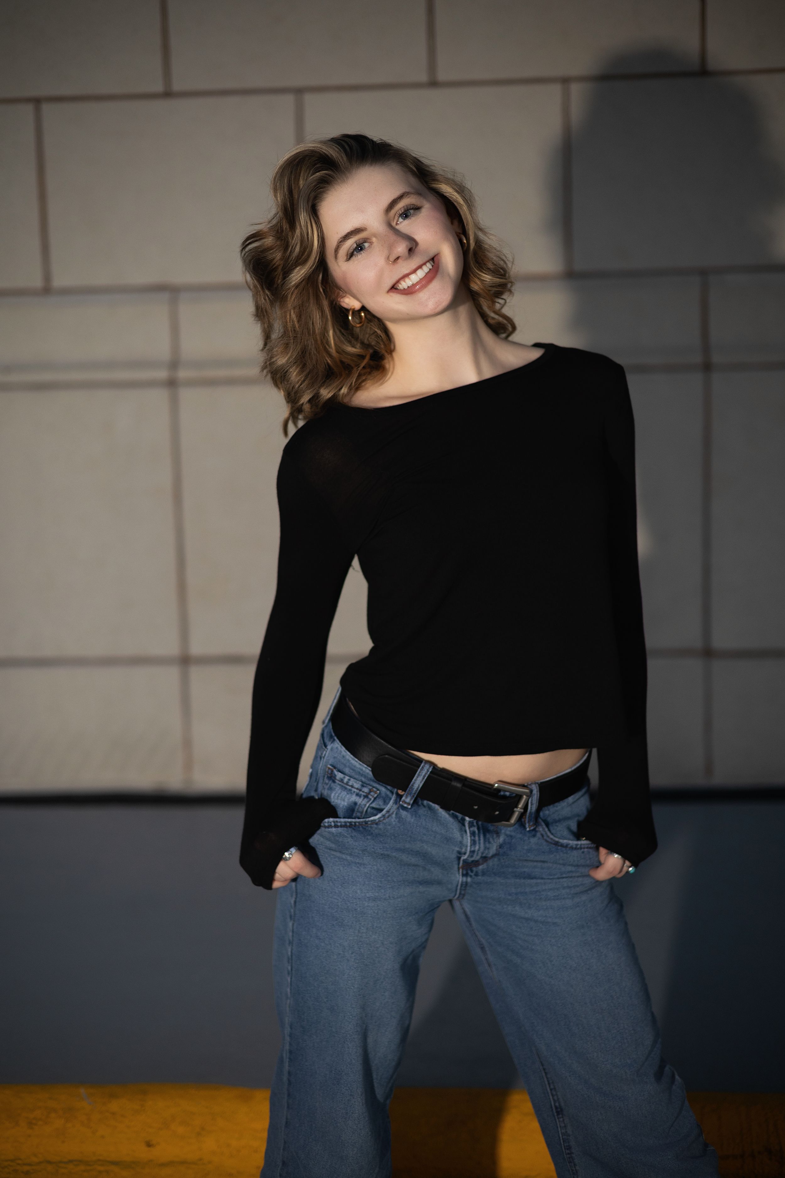 A fashion model in a black crop top and jeans poses expressively against a white wall in an industrial setting.