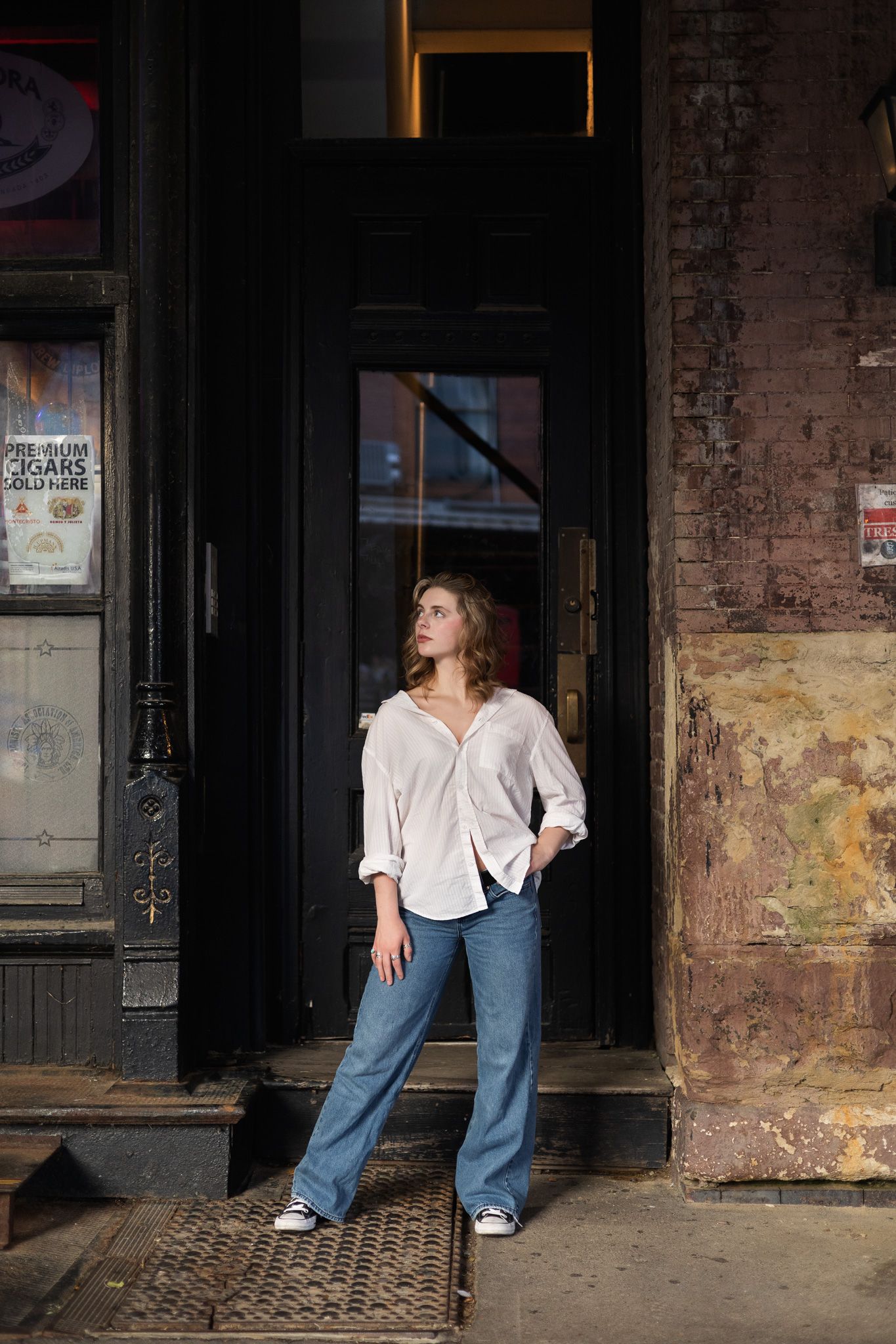 Figure in white button-down shirt and jeans poses in doorway of urban brick building.