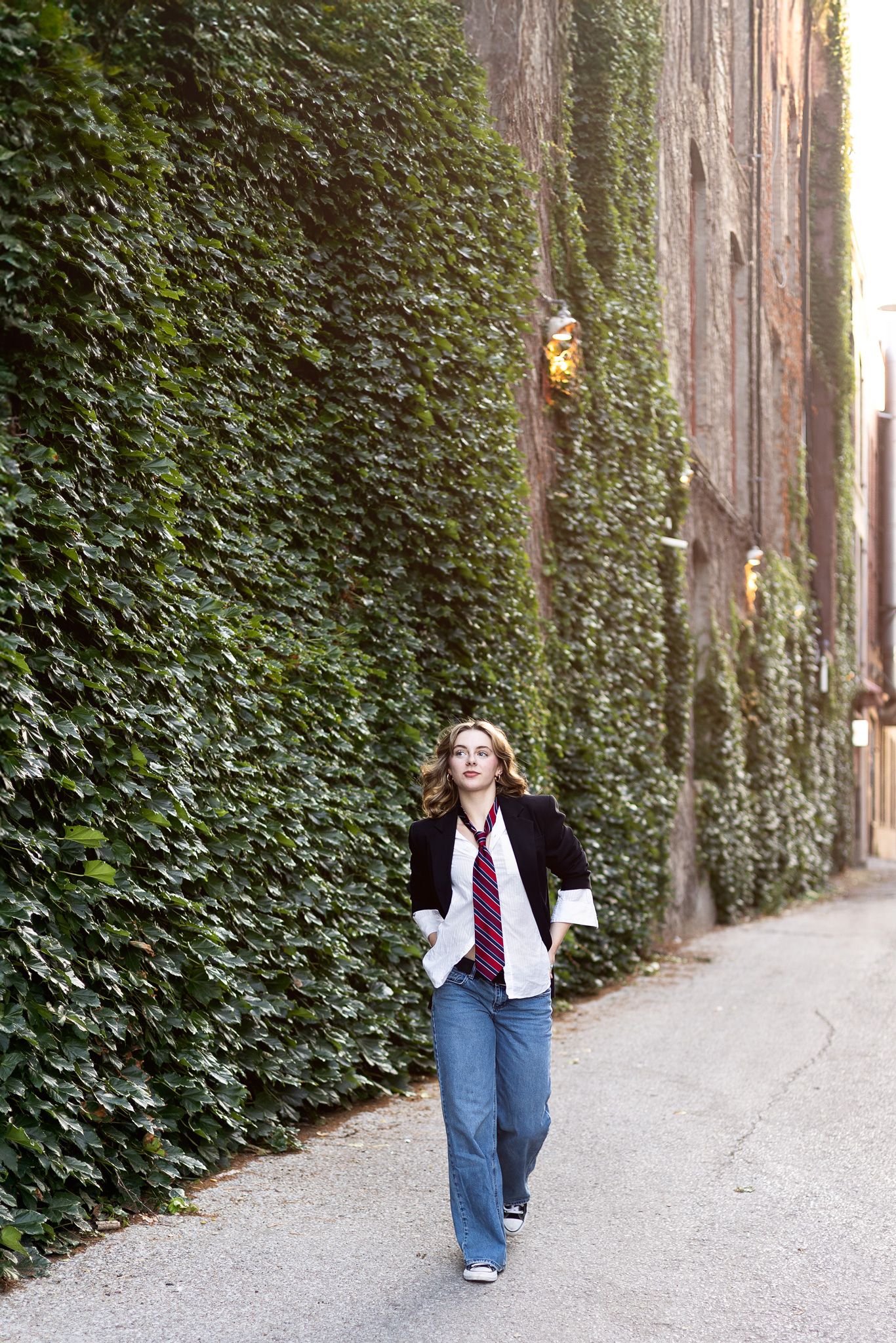 Person walking on pathway with ivy covered wall wearing black jacket and jeans in outdoor setting.