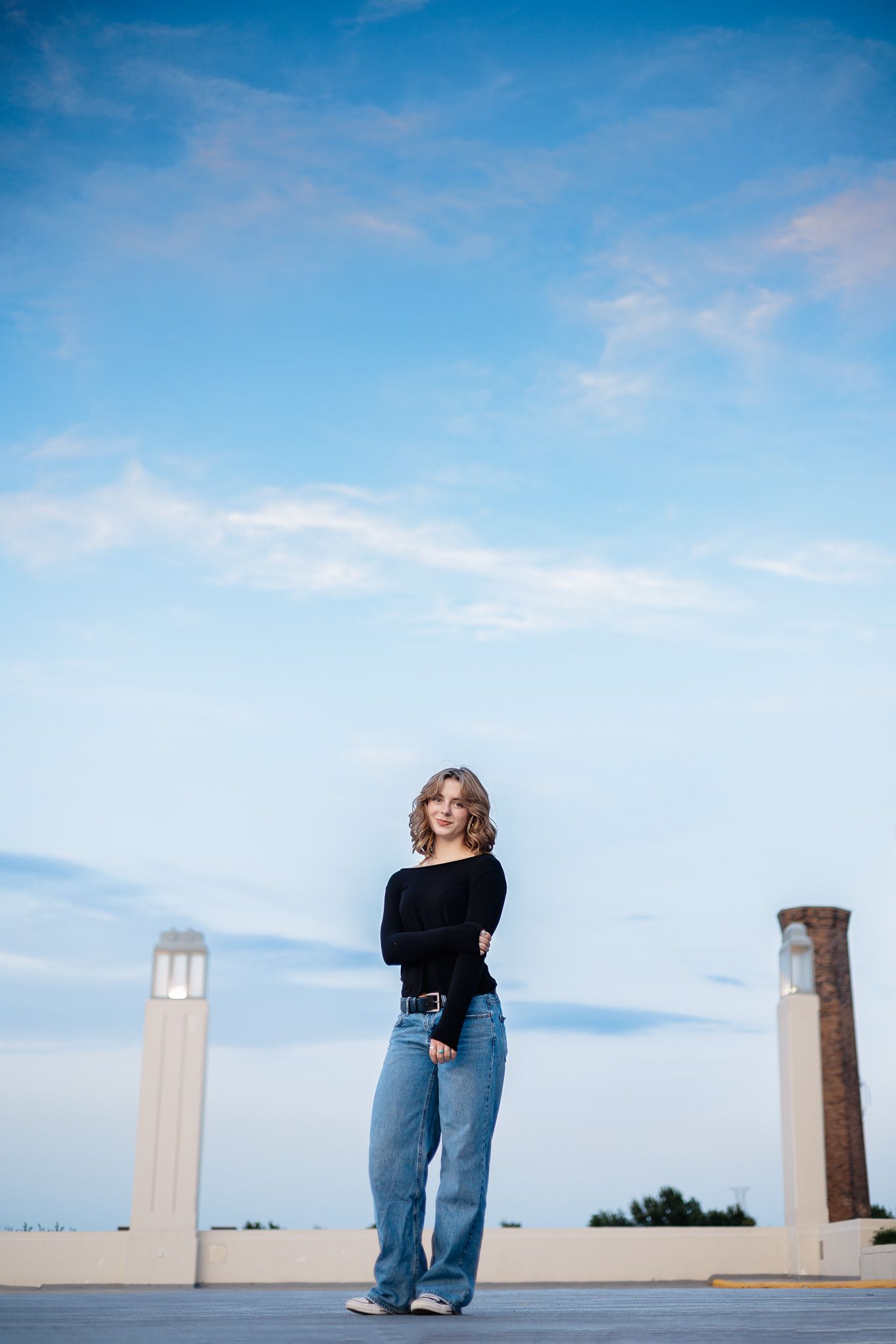 A figure in a black sweater and baggy jeans stands between two pillars on a rooftop against a bright blue sky.