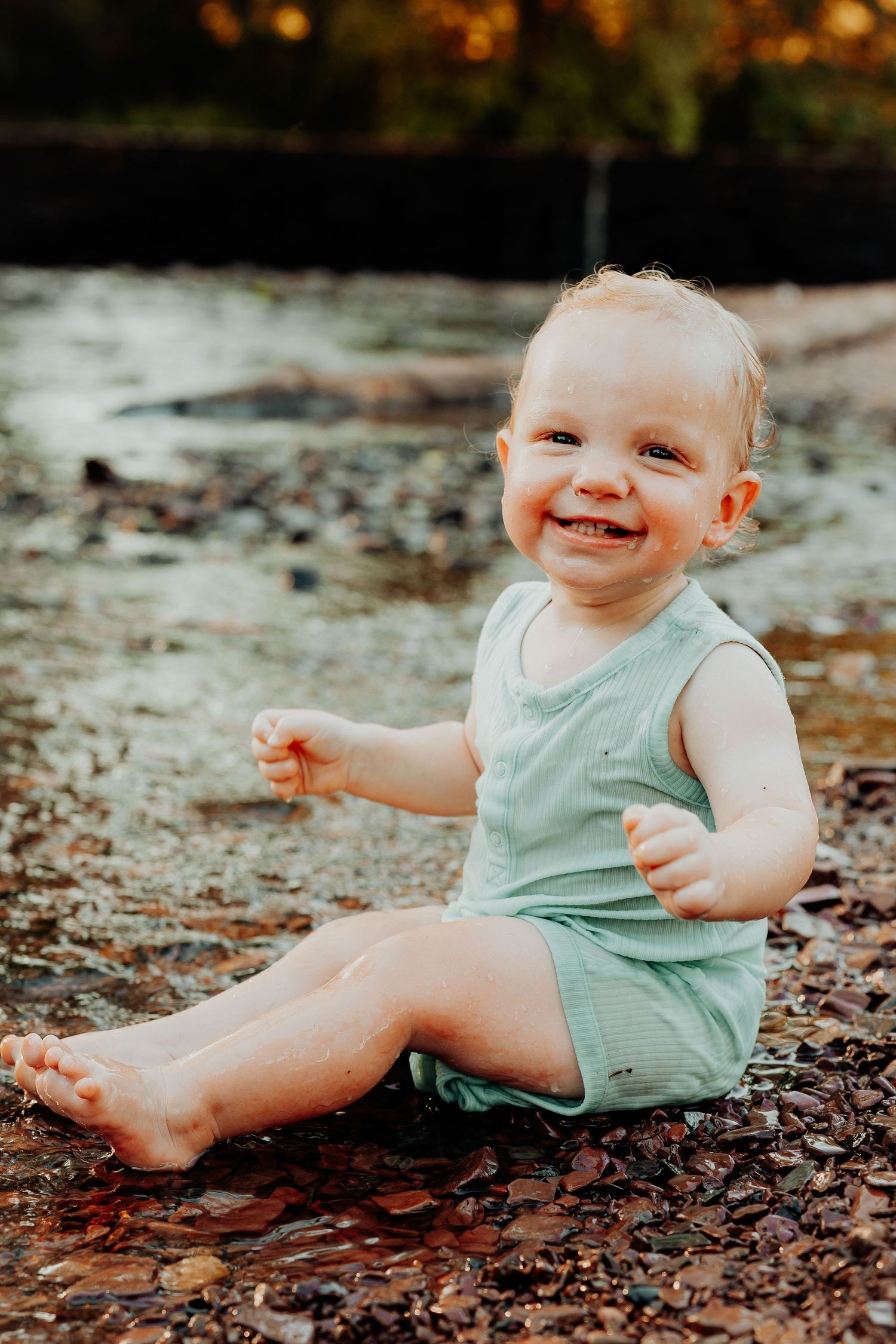 Baby wearing mint green romper sits playfully on ground covered in autumn leaves.