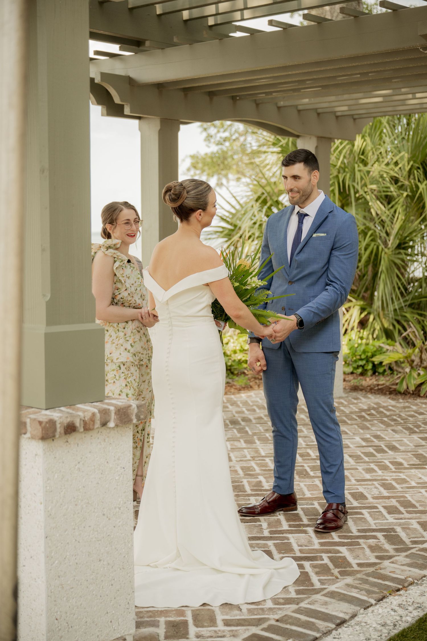 A wedding ceremony takes place under a white pergola with brick pavers and tropical landscaping in the background.
