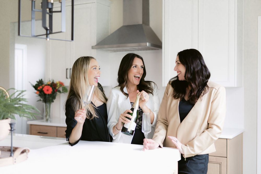 Three women laughing together while socializing and holding drinks at a kitchen counter in a modern home.