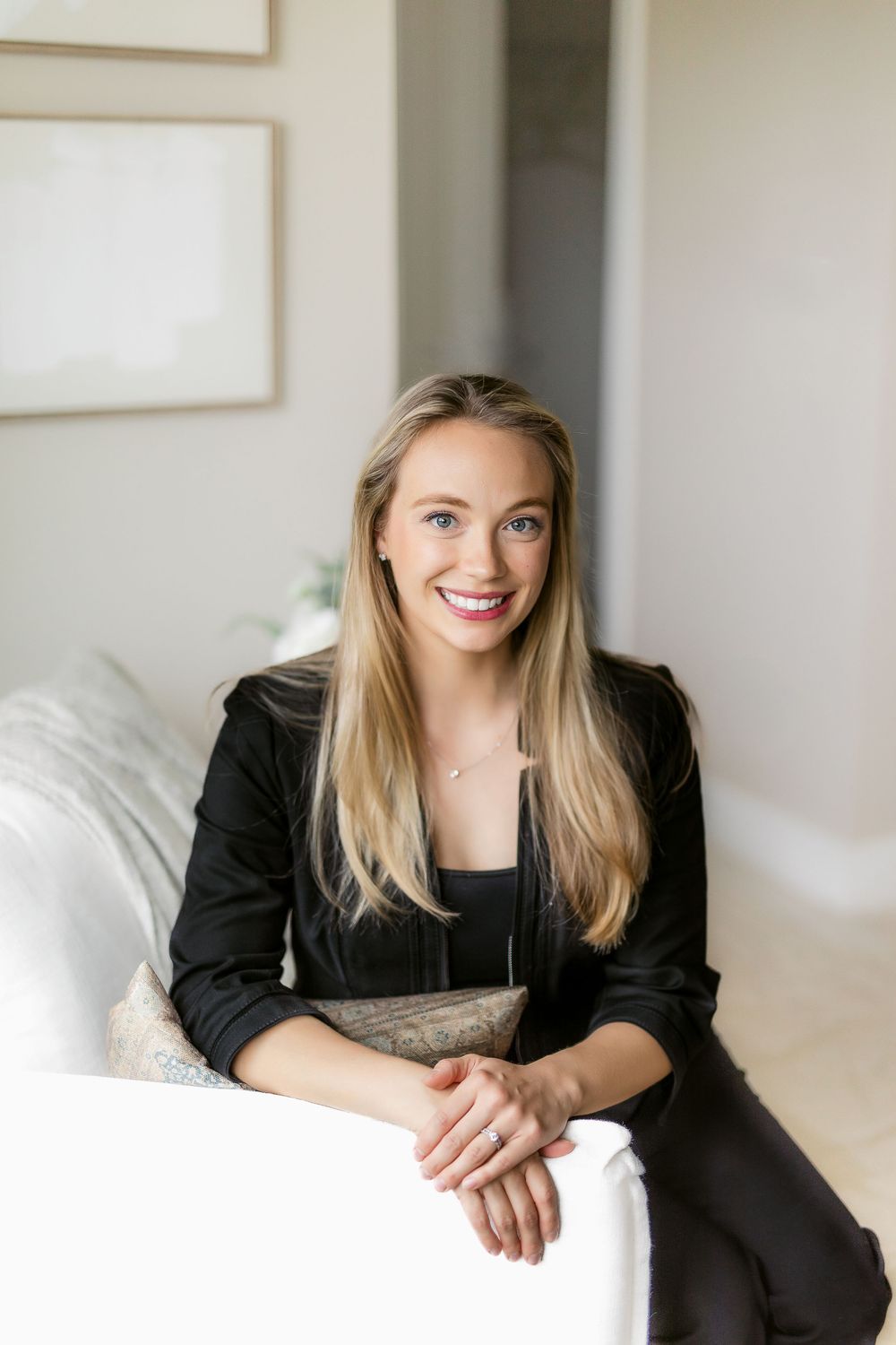 A smiling woman with long blonde hair wearing a black blazer sits casually on a white couch in a bright interior setting.
