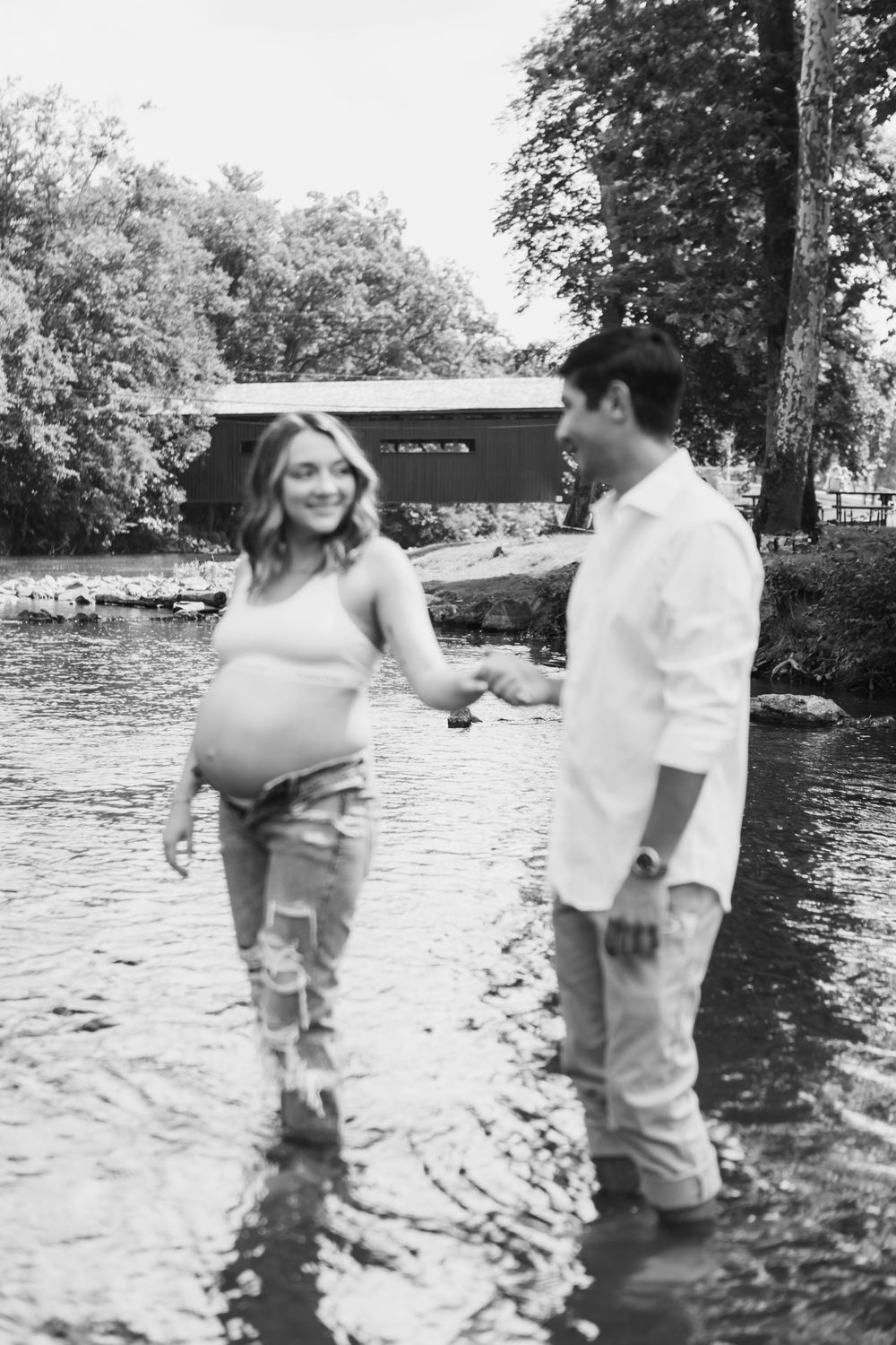 Couple stands in shallow water during black and white maternity photoshoot near covered bridge.