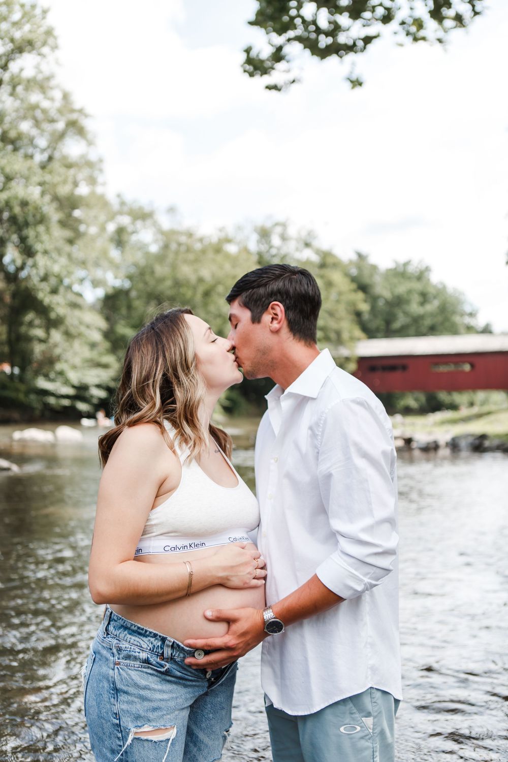 Intimate maternity photo of couple by river with covered bridge in background.