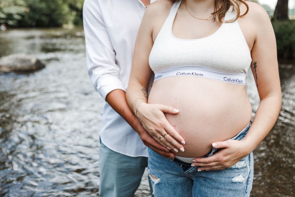 An intimate maternity photoshoot by a stream shows hands cradling a pregnant belly in casual jeans and white top.
