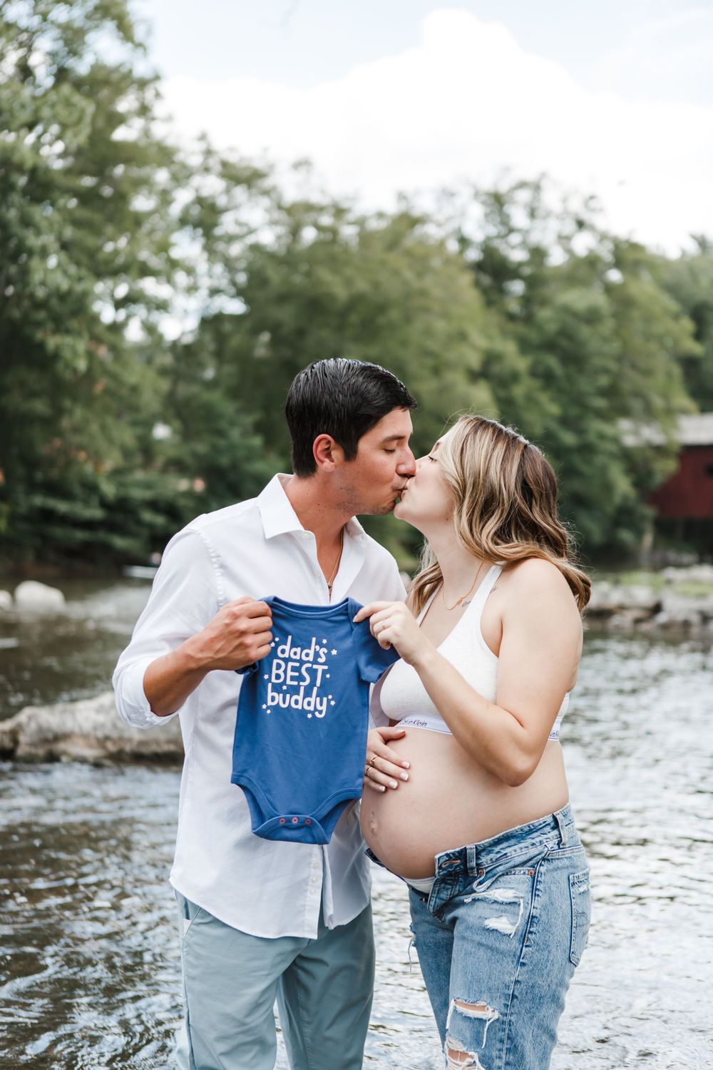 Expecting couple share romantic moment while holding baby onesie in river setting.