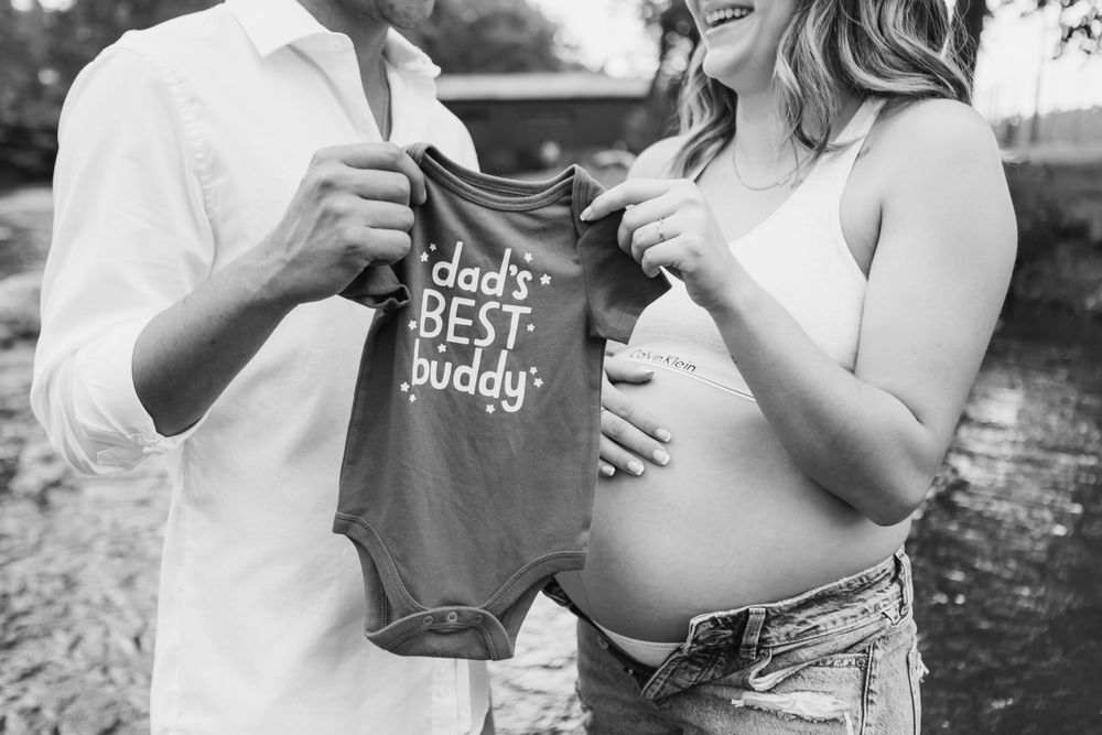 Black and white maternity photo showing pregnant woman and partner holding small baby onesie with text reading daddys best buddy.