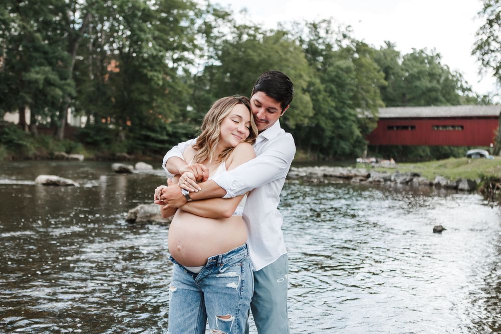 A couple embraces by a peaceful river with a red covered bridge visible in the background.