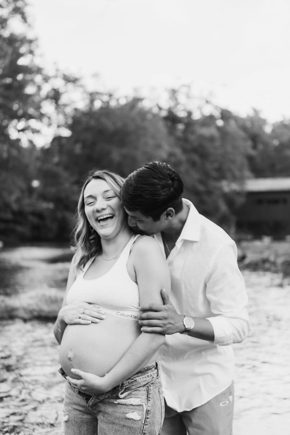 Black and white maternity photos of a couple embracing joyfully by the water's edge.