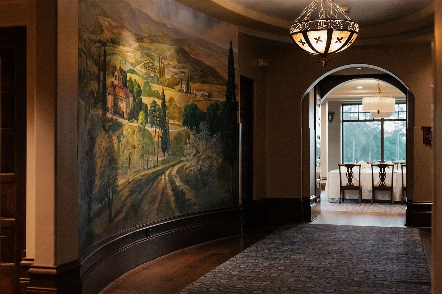 Elegant hallway with arched doorway features a large scenic countryside mural and ornate hanging light fixture.