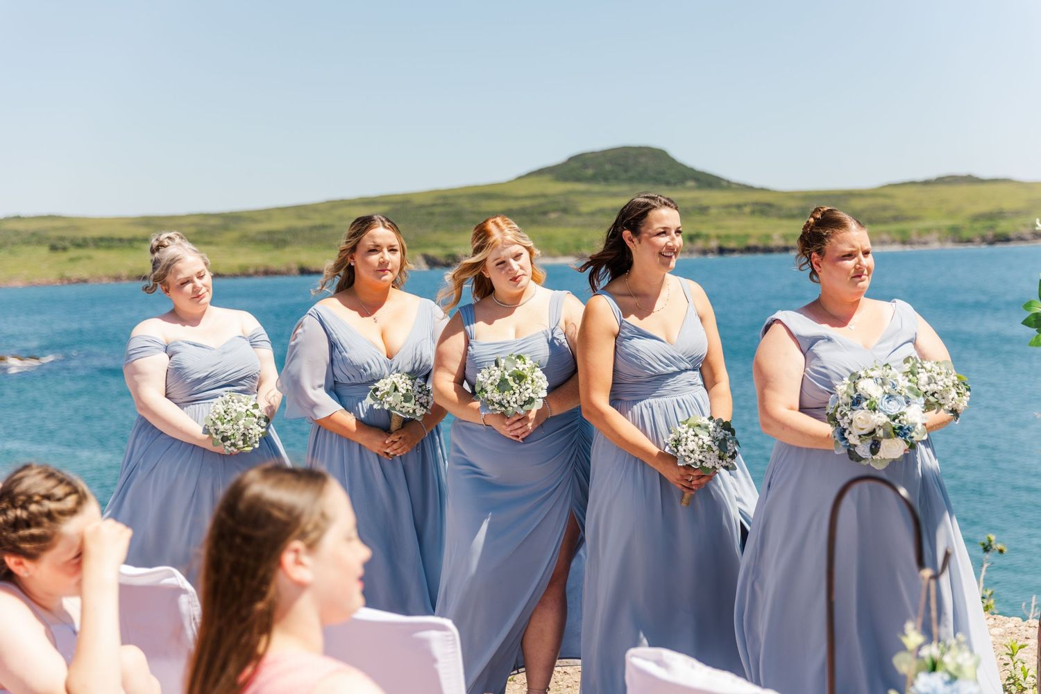 A group of bridesmaids in light blue dresses stand at an outdoor beach wedding ceremony holding white bouquets.