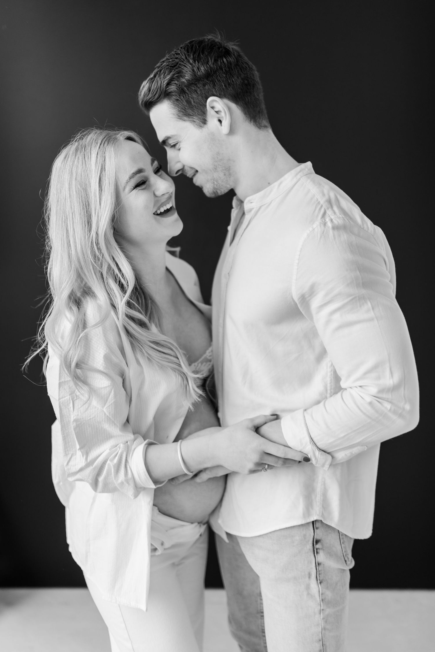 Black and white portrait of a laughing couple in white shirts sharing an intimate moment against dark background.