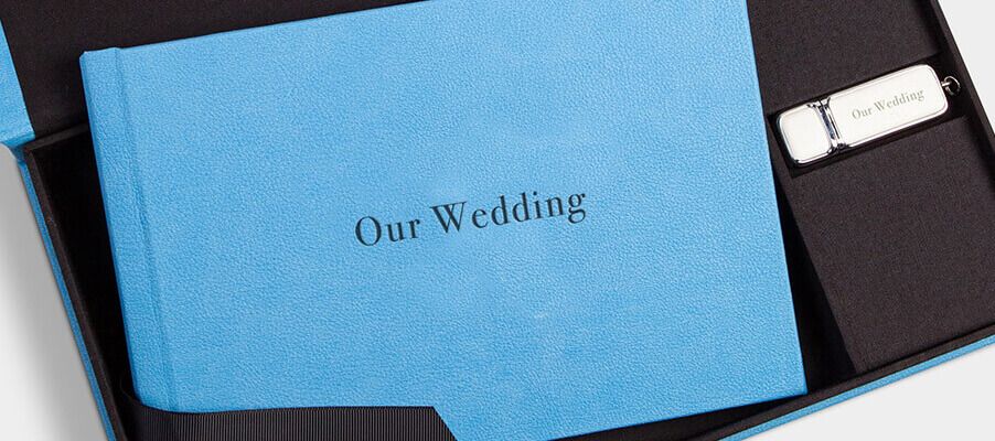 A light blue leather wedding photo album with embossed text lies surrounded by black photo accessories.